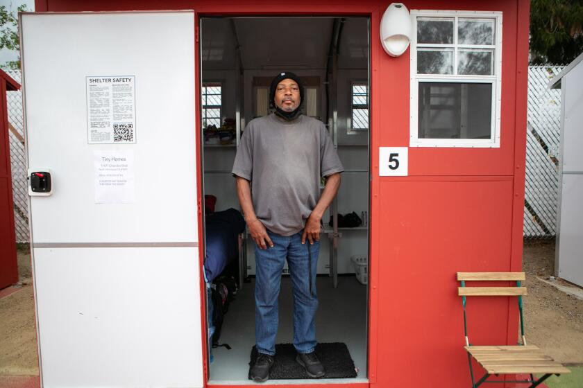 Stephen Smith, in grey shirt and jeans, stands in the doorway of a tiny home that is painted red
