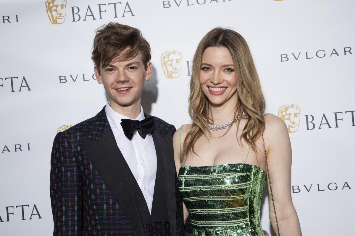 Thomas Brodie-Sangster in a plaid tuxedo jacket posing with Talulah Riley in a green-sequined dress