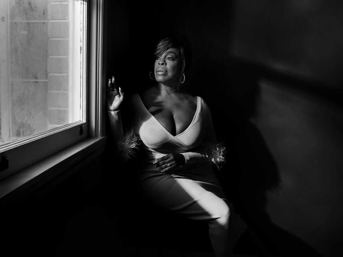 In a black and white portrait, Niecy Nash-Betts sits in the shadows by a corner window. 