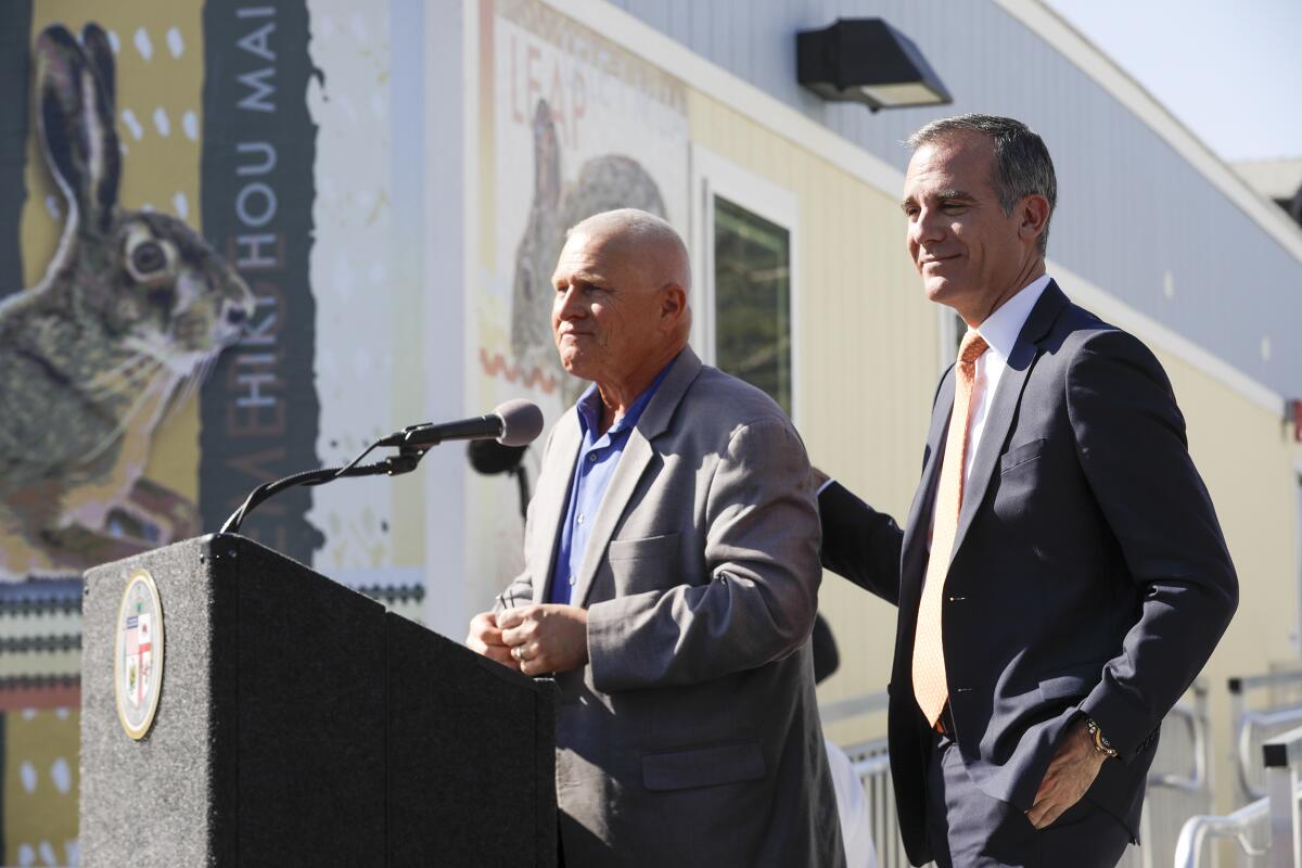 Mike Bonin speaks into a microphone. Eric Garcetti stands behind him with one hand on Bonin's back.