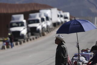 A street vendor in a white hat stands under a blue umbrella at his stand, across the road from a line of white trucks
