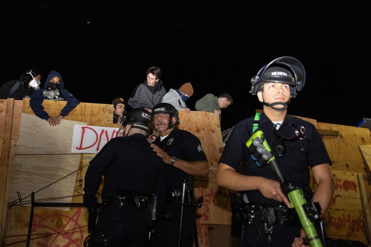Demonstrators stand behind plywood as authorities stand below the structure and one holds a long yellow and black weapon.