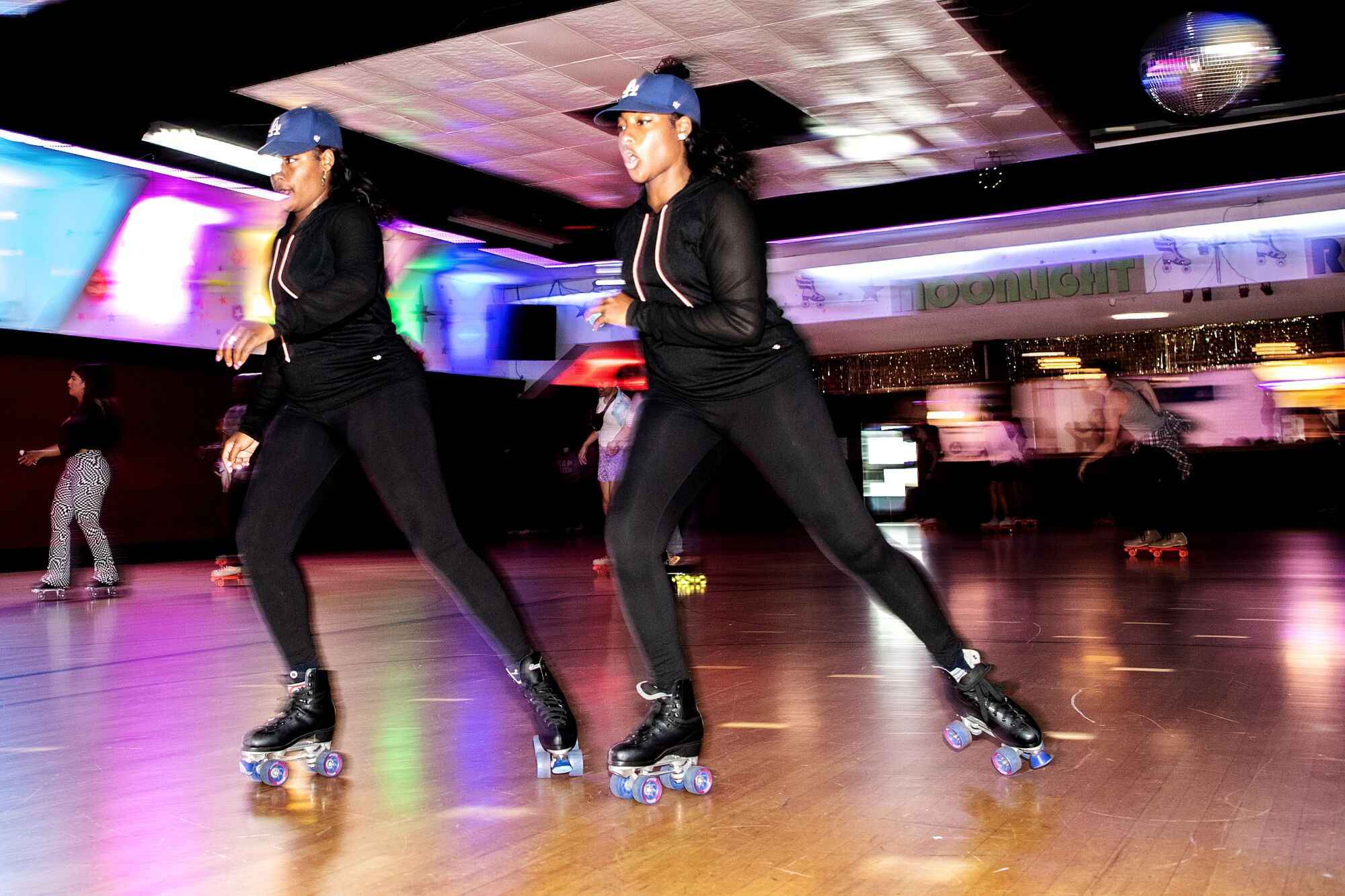 Two people wearing the same all-black outfit and blue caps while skating.