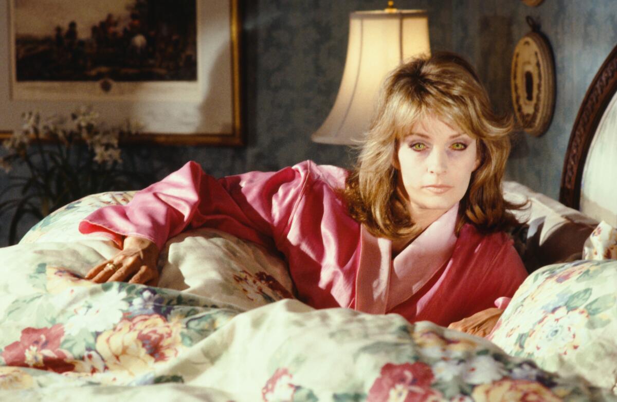 A yellow-eyed Deidre Hall lies in bed in a pink bathrobe.
