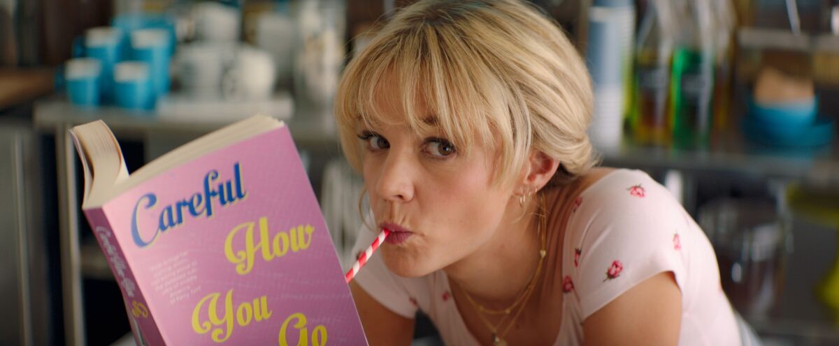 Carey Mulligan drinks from a straw while reading a book in a scene in "Promising Young Woman." 