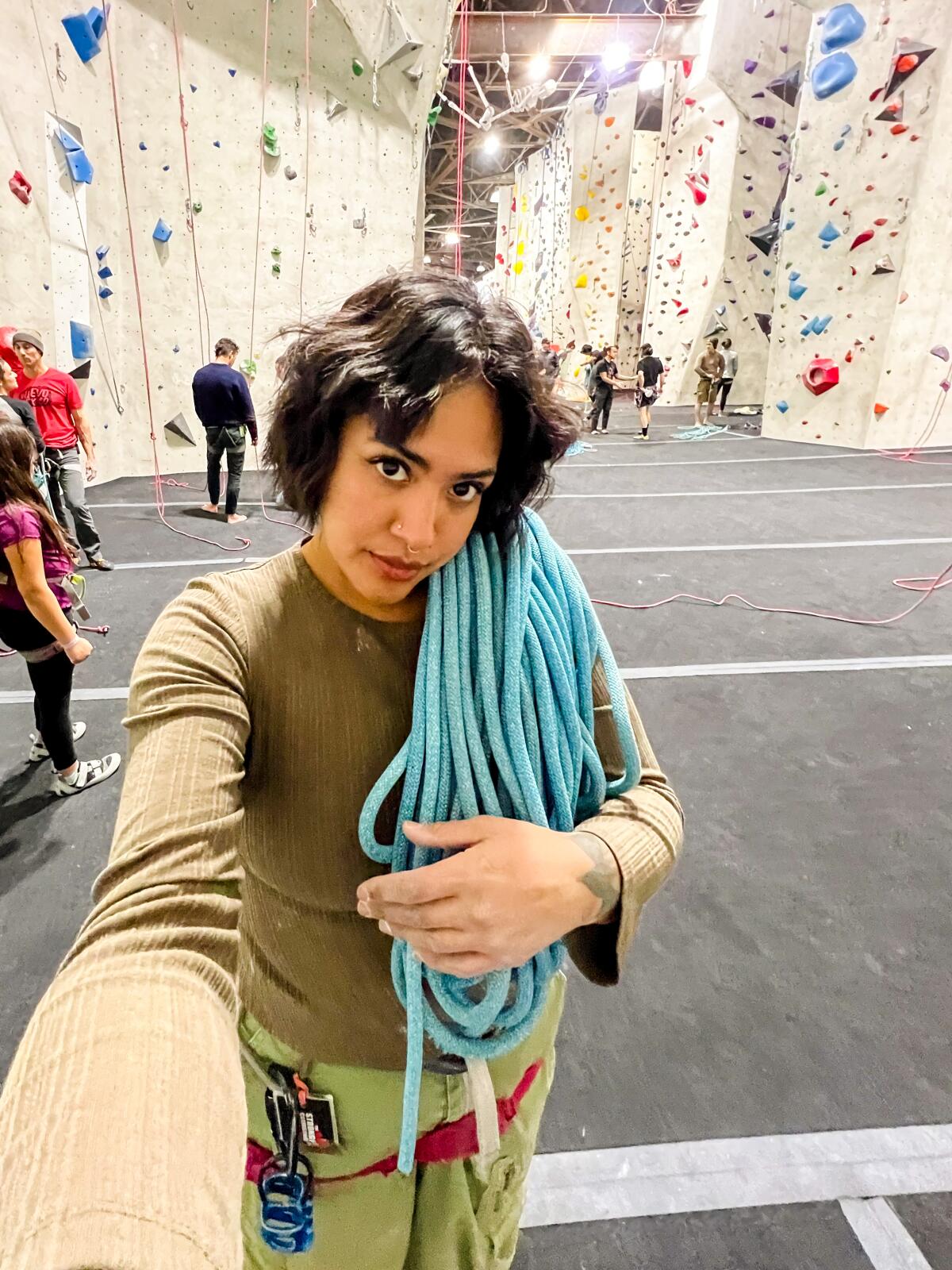 A selfie of Yanira Fuentes at Stronghold Climbing Gym with climbing walls behind her and rope over her shoulder