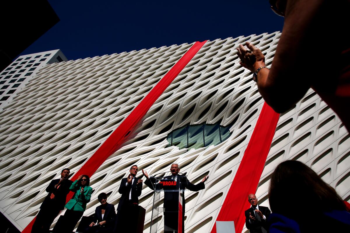 Eli Broad stands with arms outstretched at a lectern in front of a white contemporary art museum with two large red ribbons