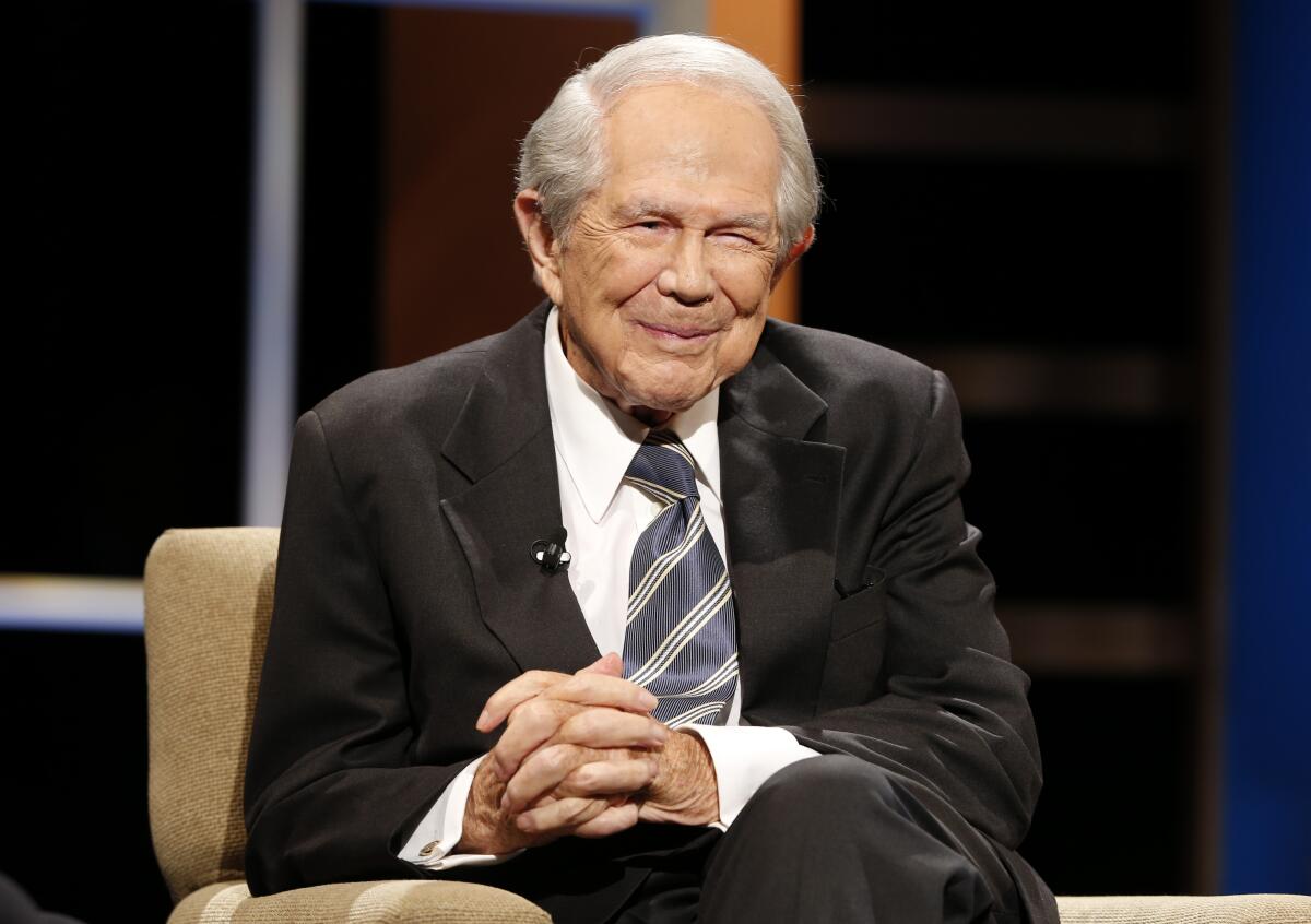 Rev. Pat Robertson sits in a an upholstered chair on a stage, slightly smiling.