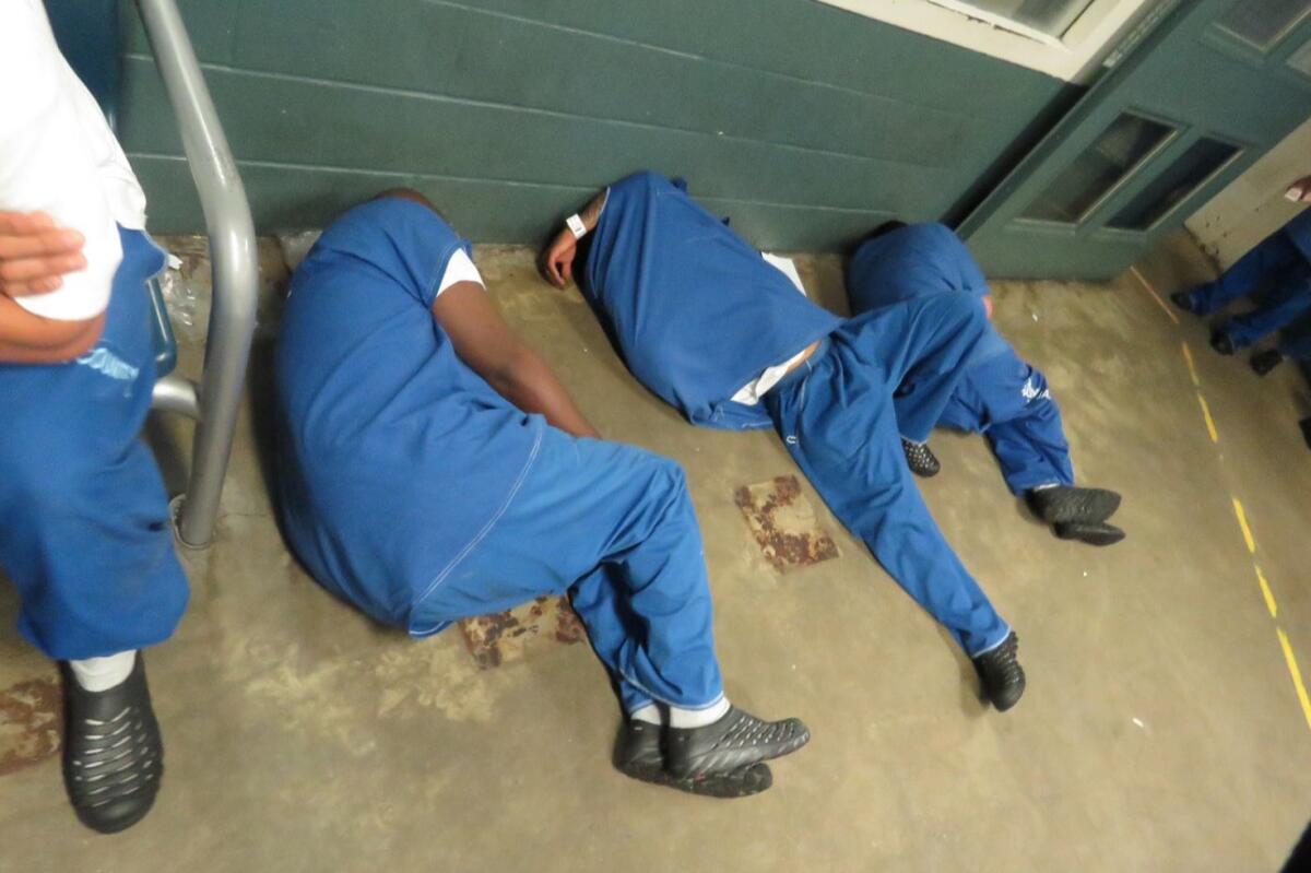 Three people dressed in blue jail clothing sleep on a bare concrete floor.