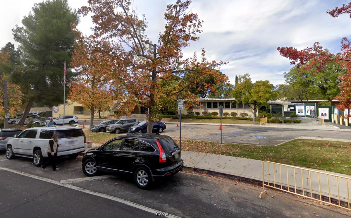 Several cars stopped on the street or parked in the lot outside a low-profile building surrounded by trees and shrubs