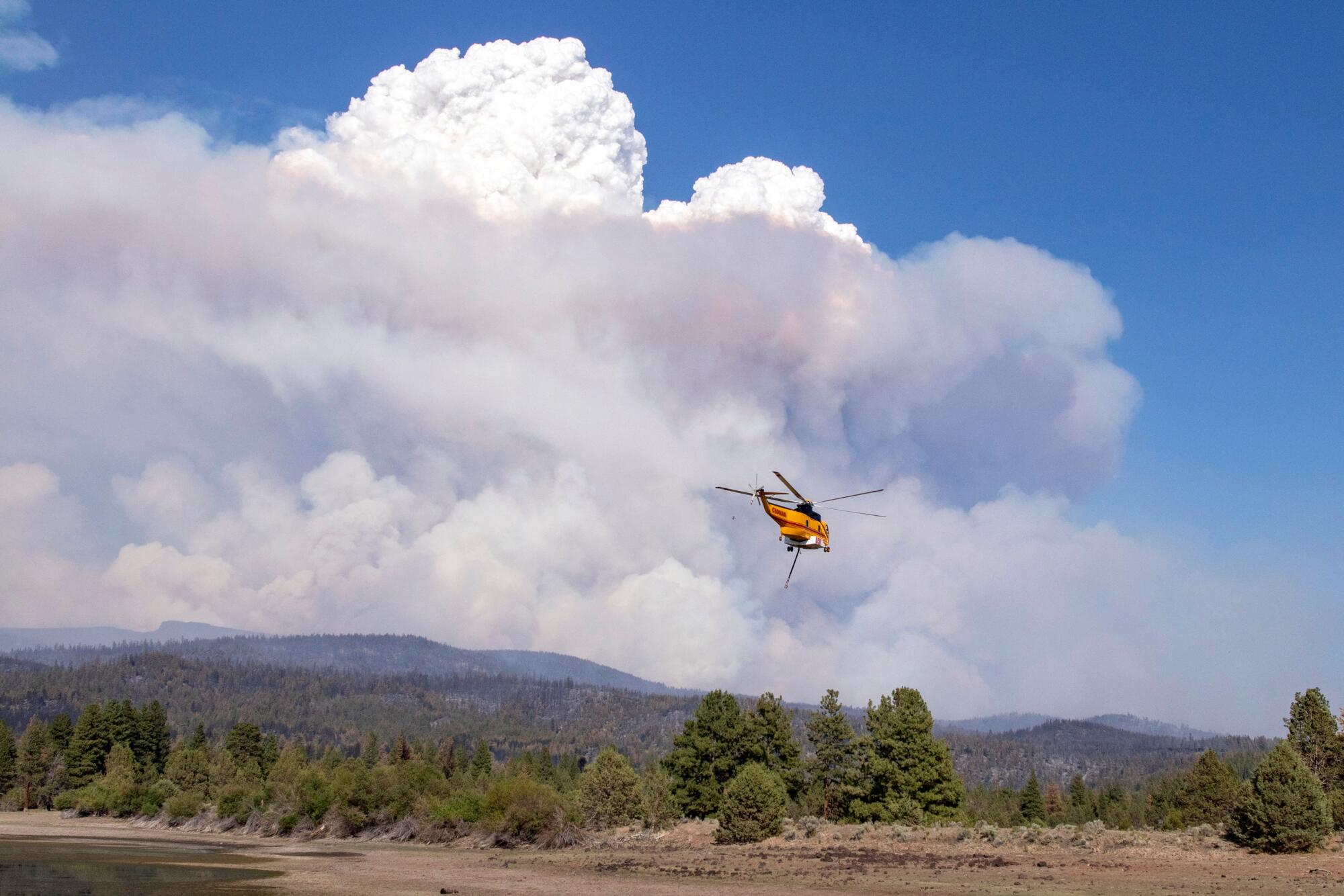 A helicopter flies with towering smoke plumes on the horizon.