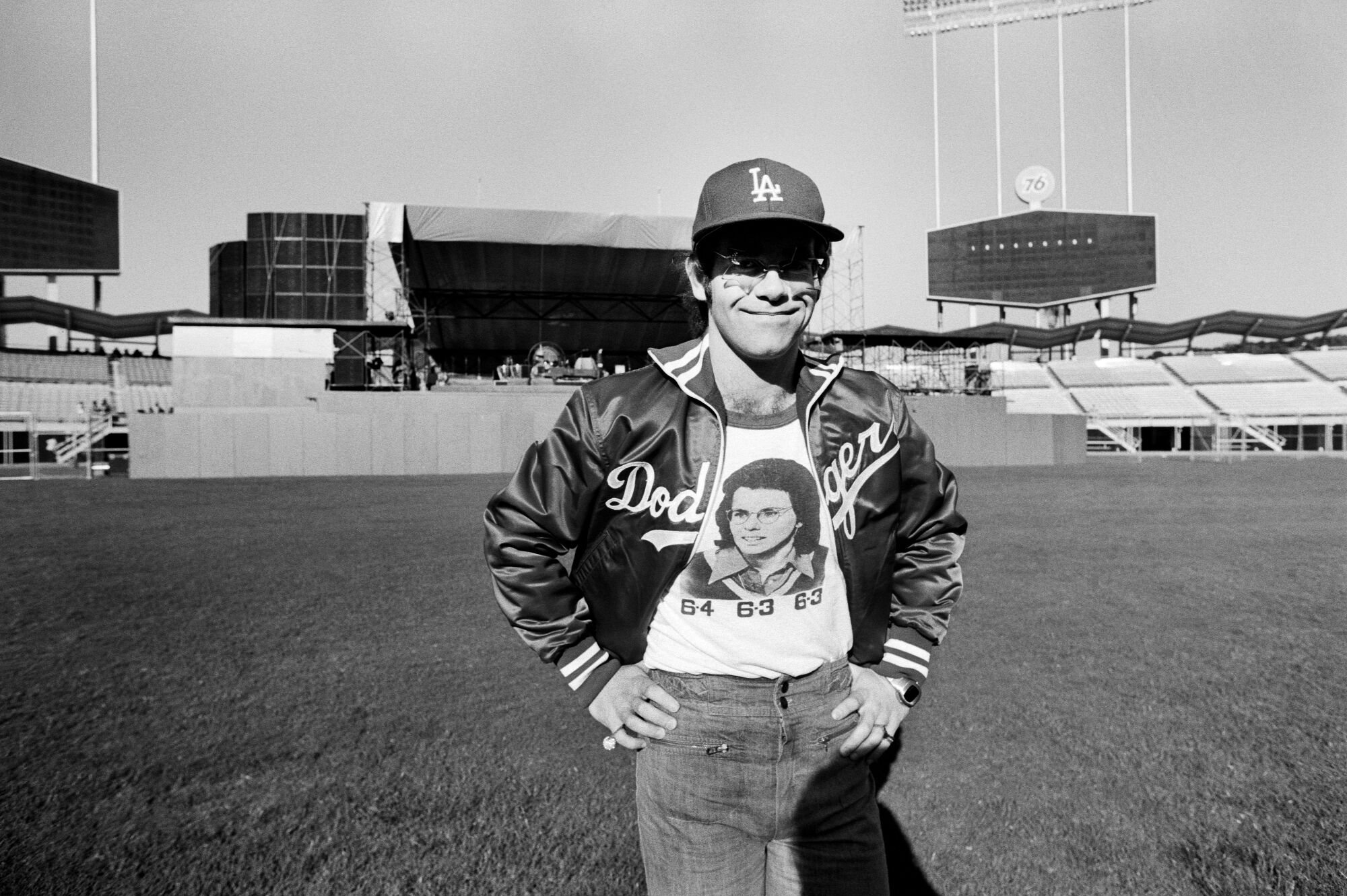 A man in an LA Dodgers cap and jacket stands on an empty stadium baseball field.