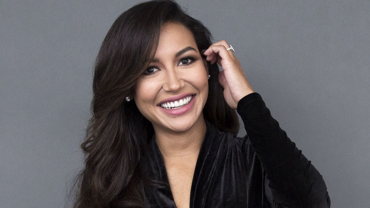 Naya Rivera in a black shirt, brushing her hair back with one hand