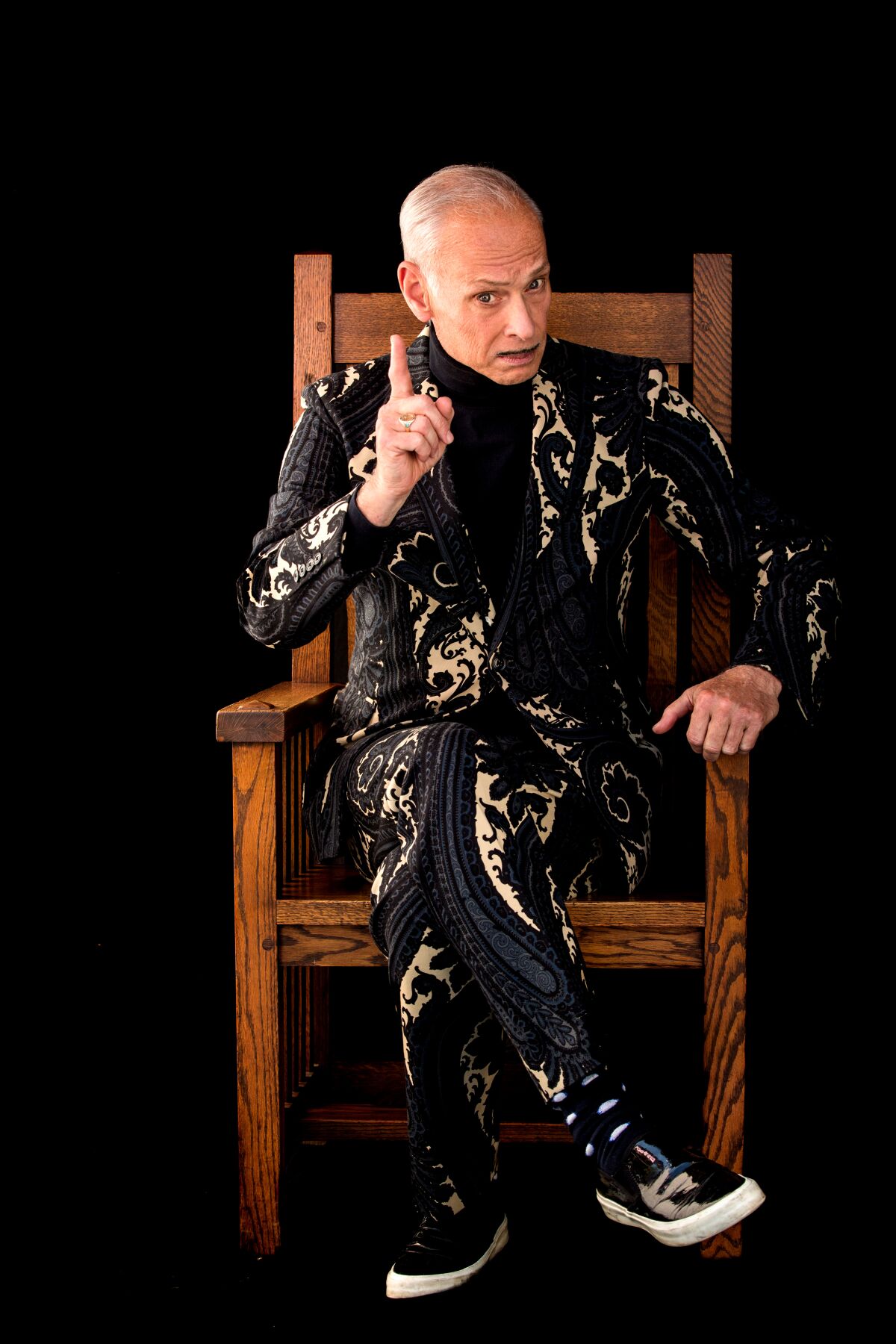 man in patterned suit sits in large wooden chair and wags his finger 