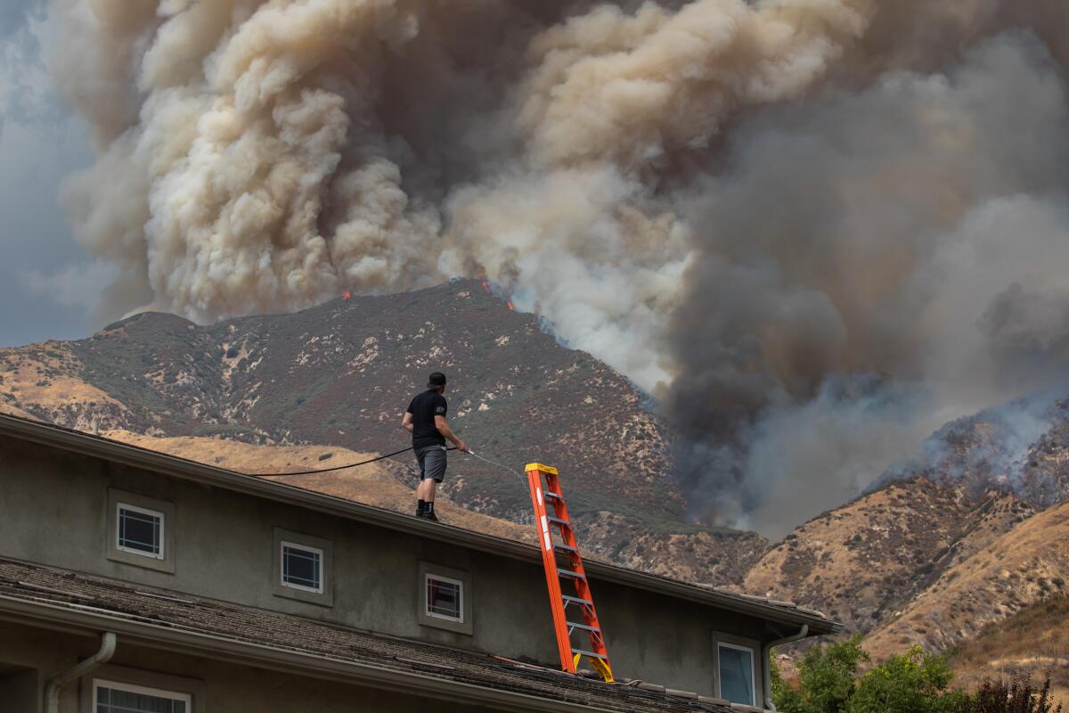 A man standing on a rooftop with a hose looks toward a massive cloud of smoke rising from a hillside.