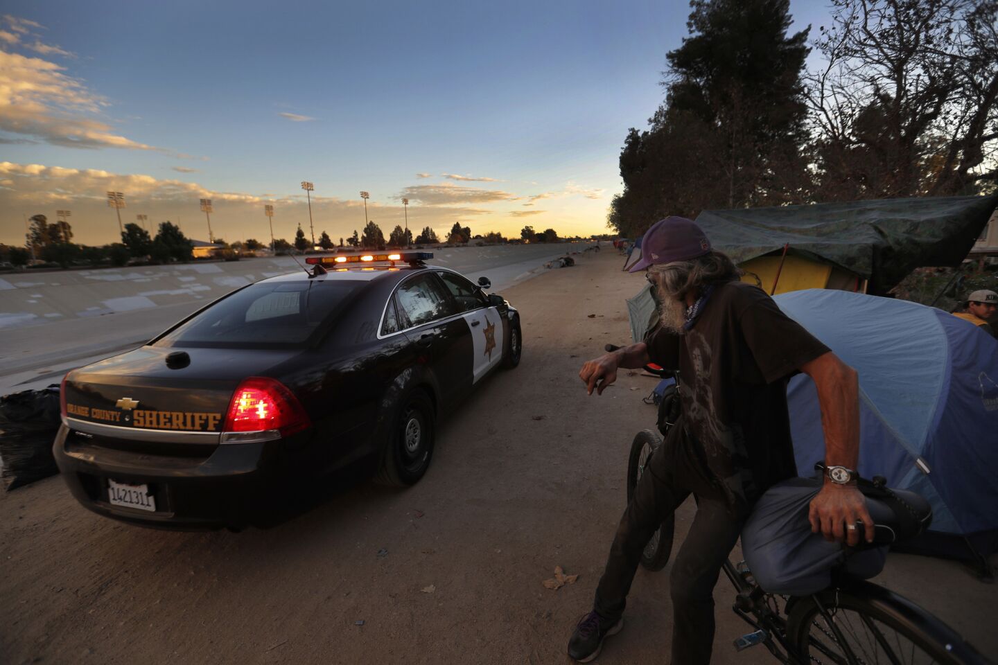 An Orange County sheriff’s cruiser drives along the Santa Ana River, past homeless encampments.