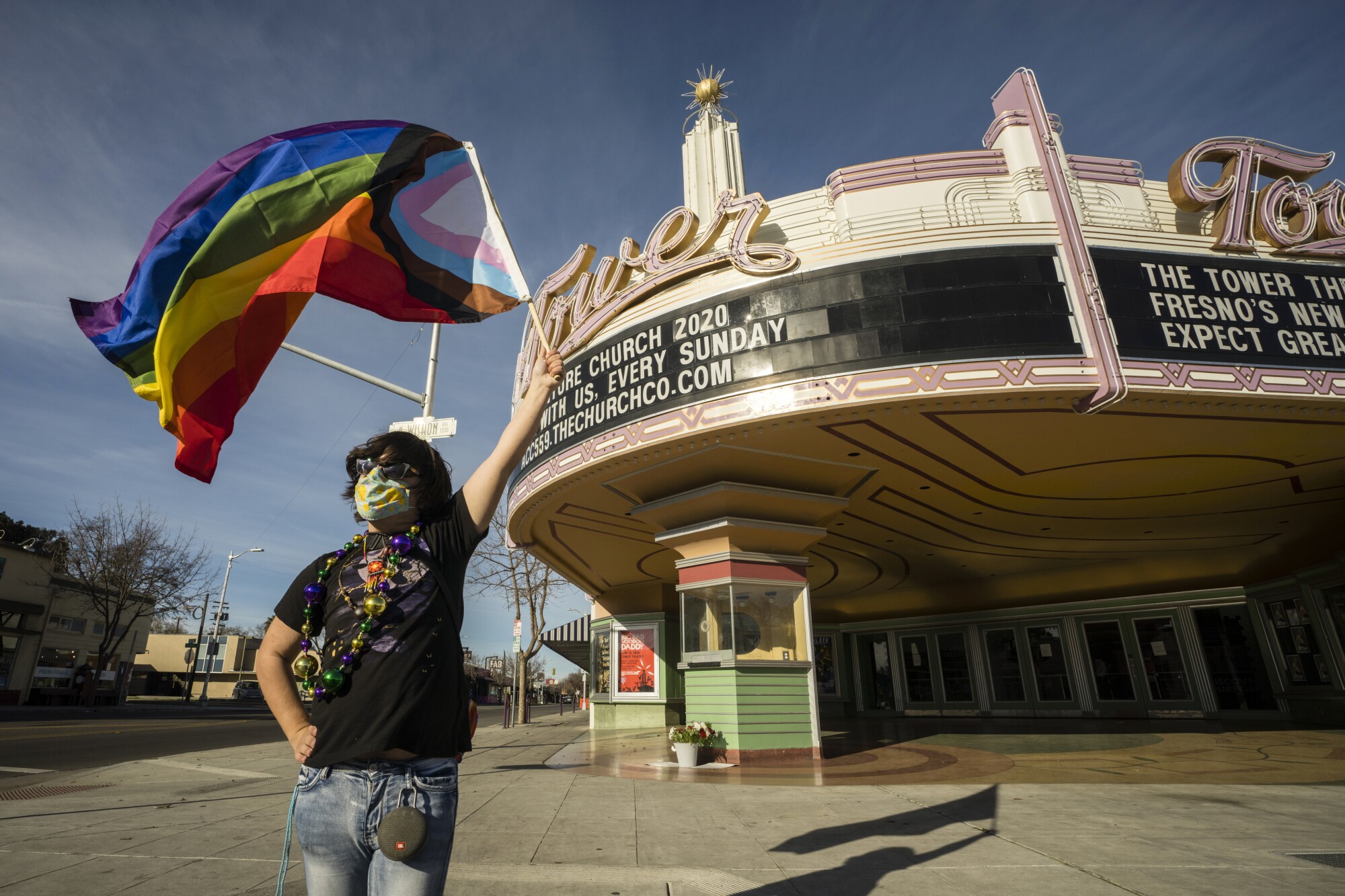 A person in a mask waves a rainbow flag in front of an Art Deco theater