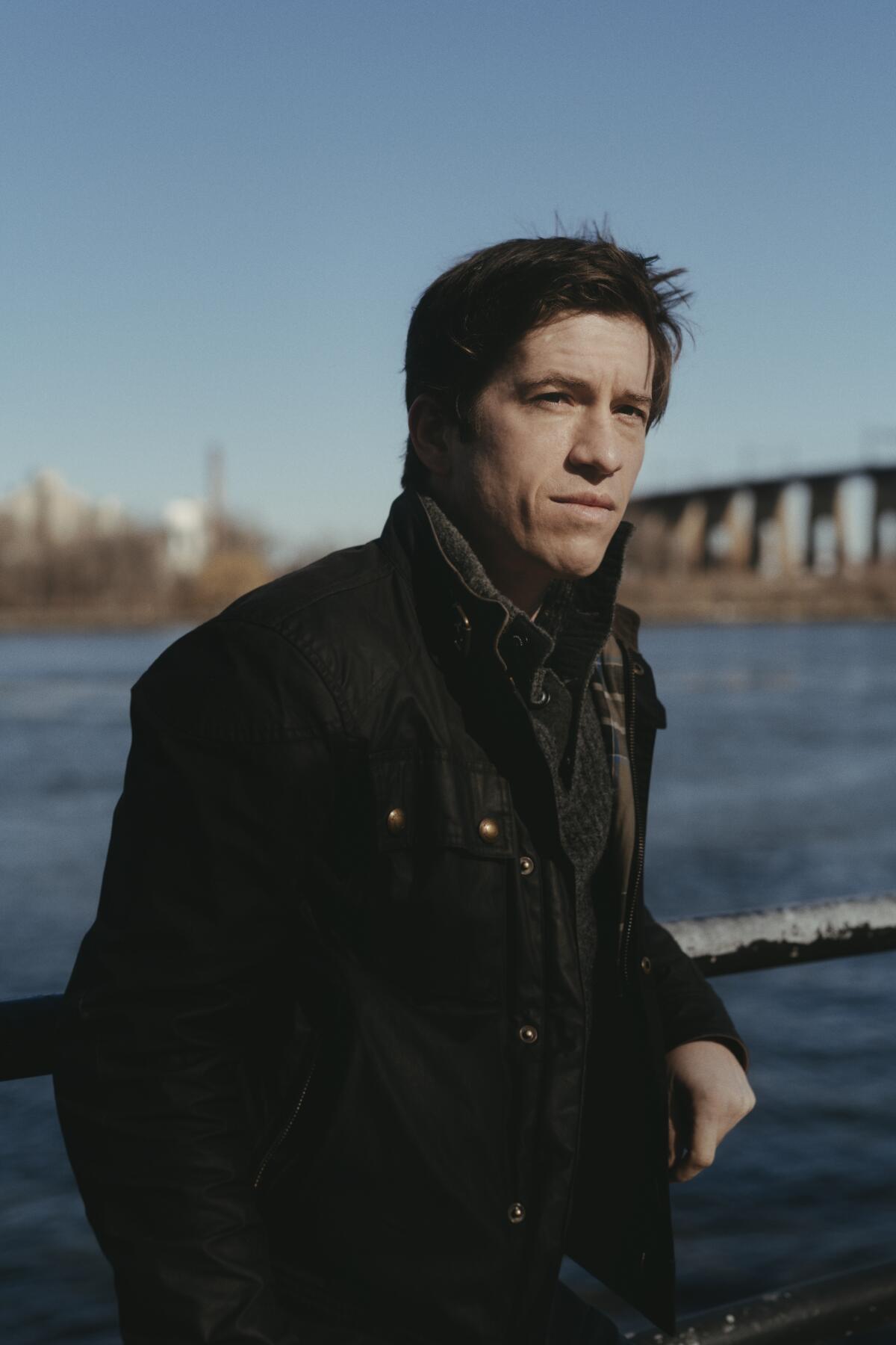 A man in a scarf and overcoat stands beside a river.