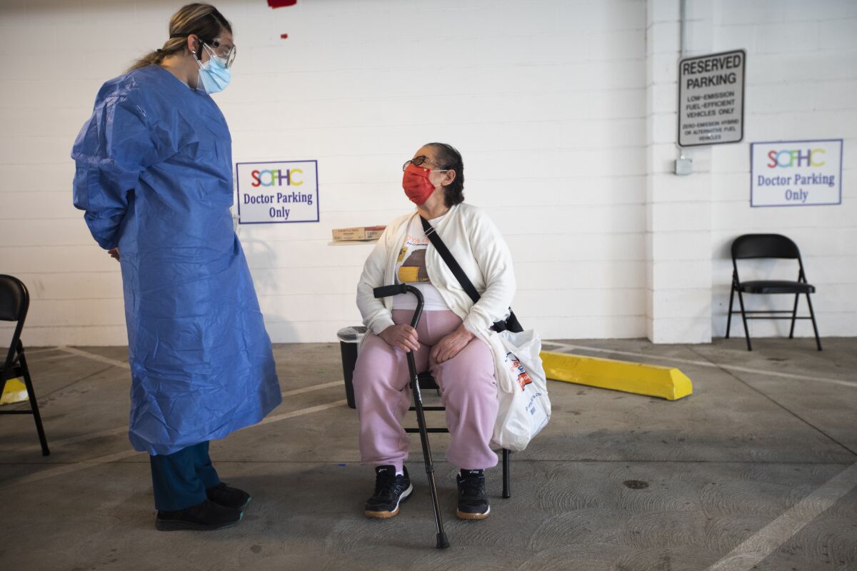 A healthcare worker in a mask and blue scrubs talks to a seated patient in an area set up in a parking garage