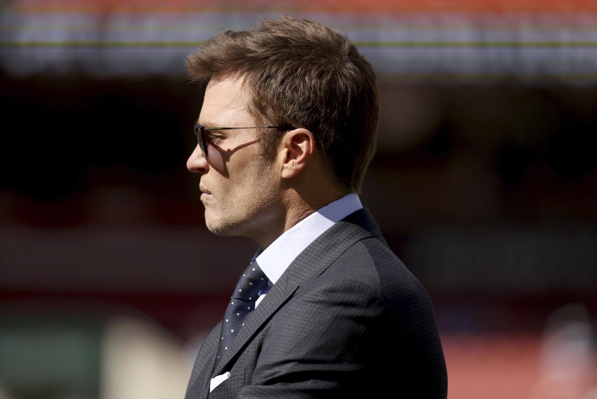 Tom Brady wears a suit, sunglasses and a serious expression in a profile shot from the football field before a Fox broadcast