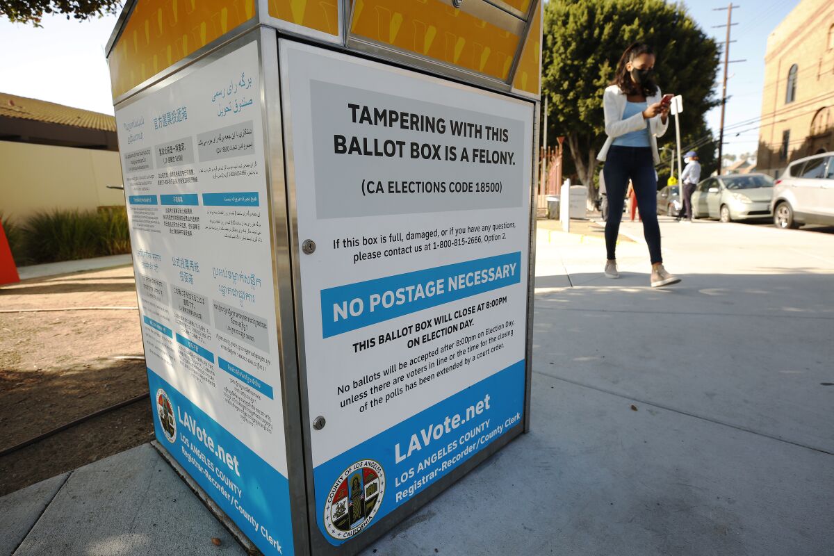 A ballot box on a city sidewalk. A woman in a mask is in the background.