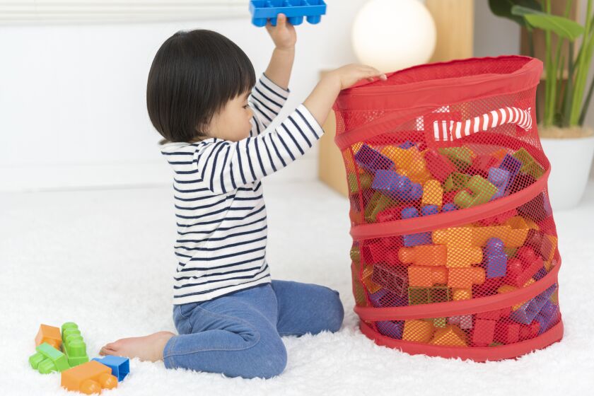 A 2-year-old girl helps clean up the room by putting toys in a basket.