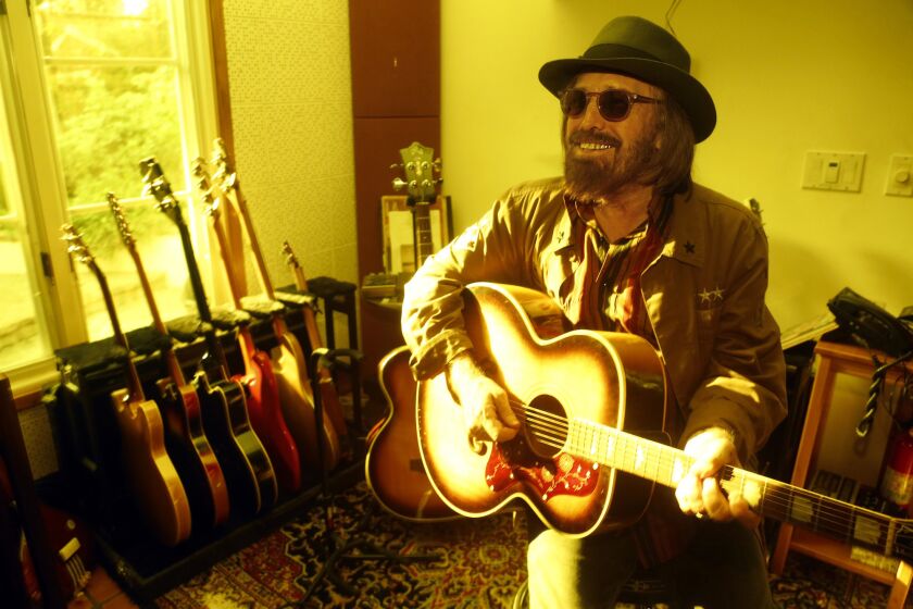 Tom Petty, dressed in a jacket, sunglasses and fedora,  smiles and holds a guitar while seated in his home.