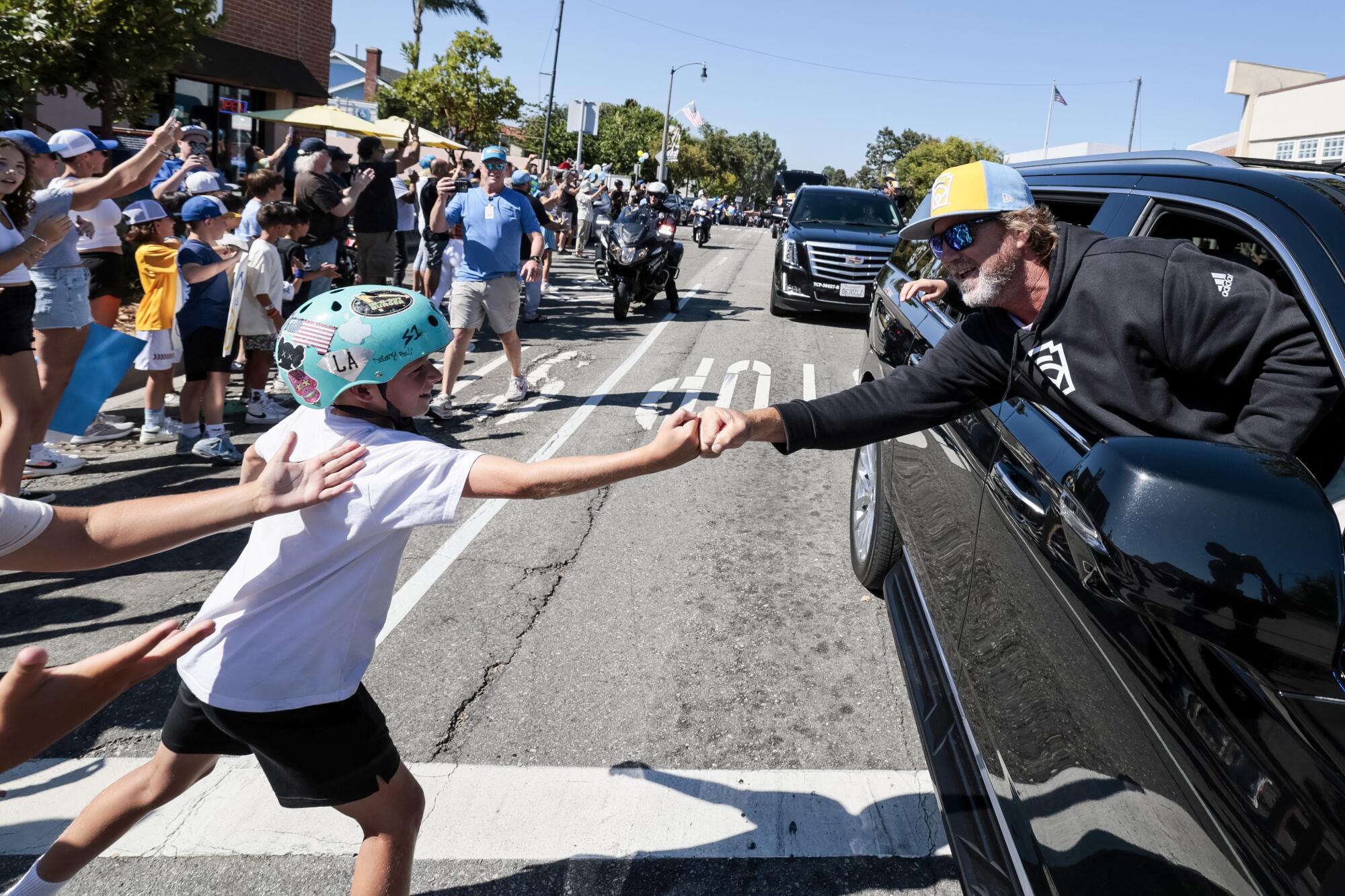 A man with a graying beard and baseball cap reaches out of a car window to clasp hands with a boy among a crowd.