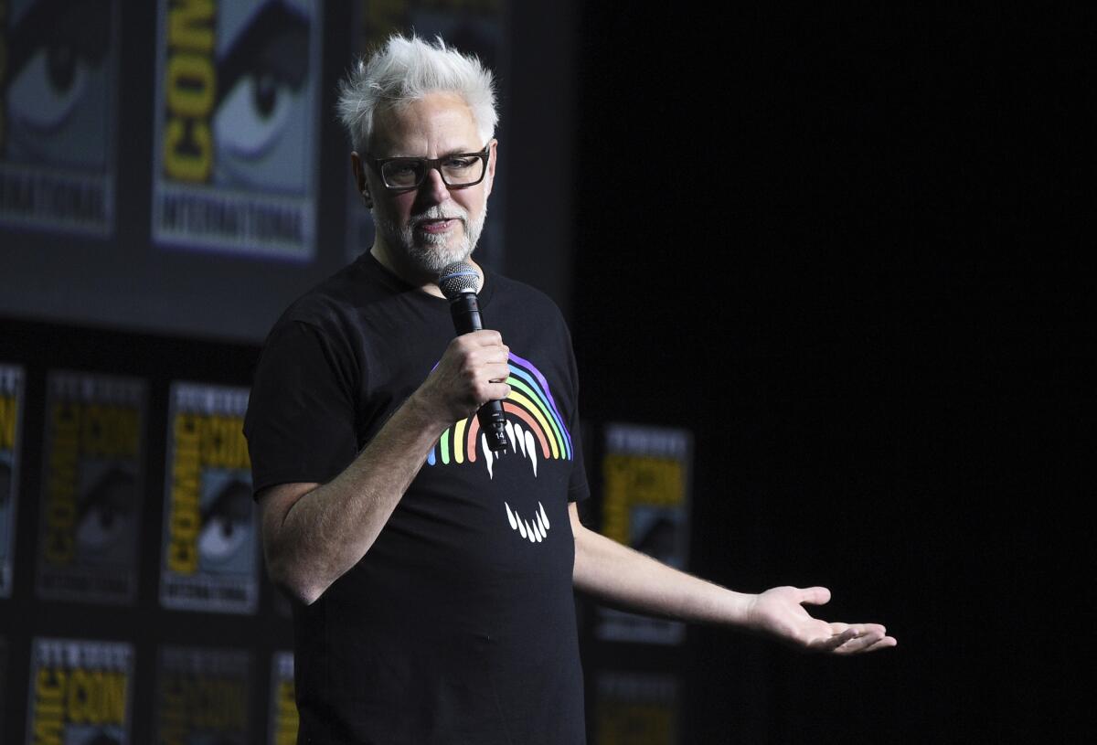 A man with white hair and a beard wearing glasses and a rainbow shirt speaks into a microphone on a stage.