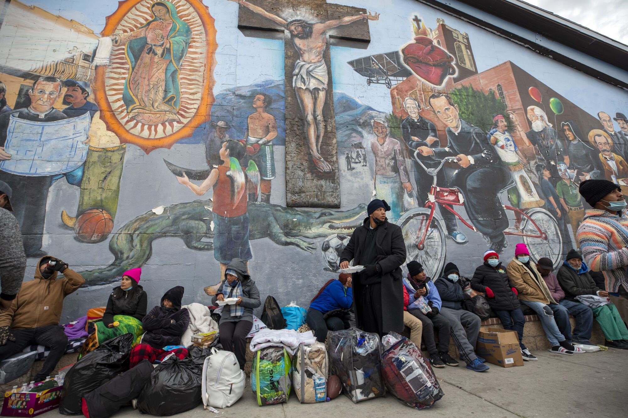 People with baggage sit on the sidewalk outside beneath a mural 