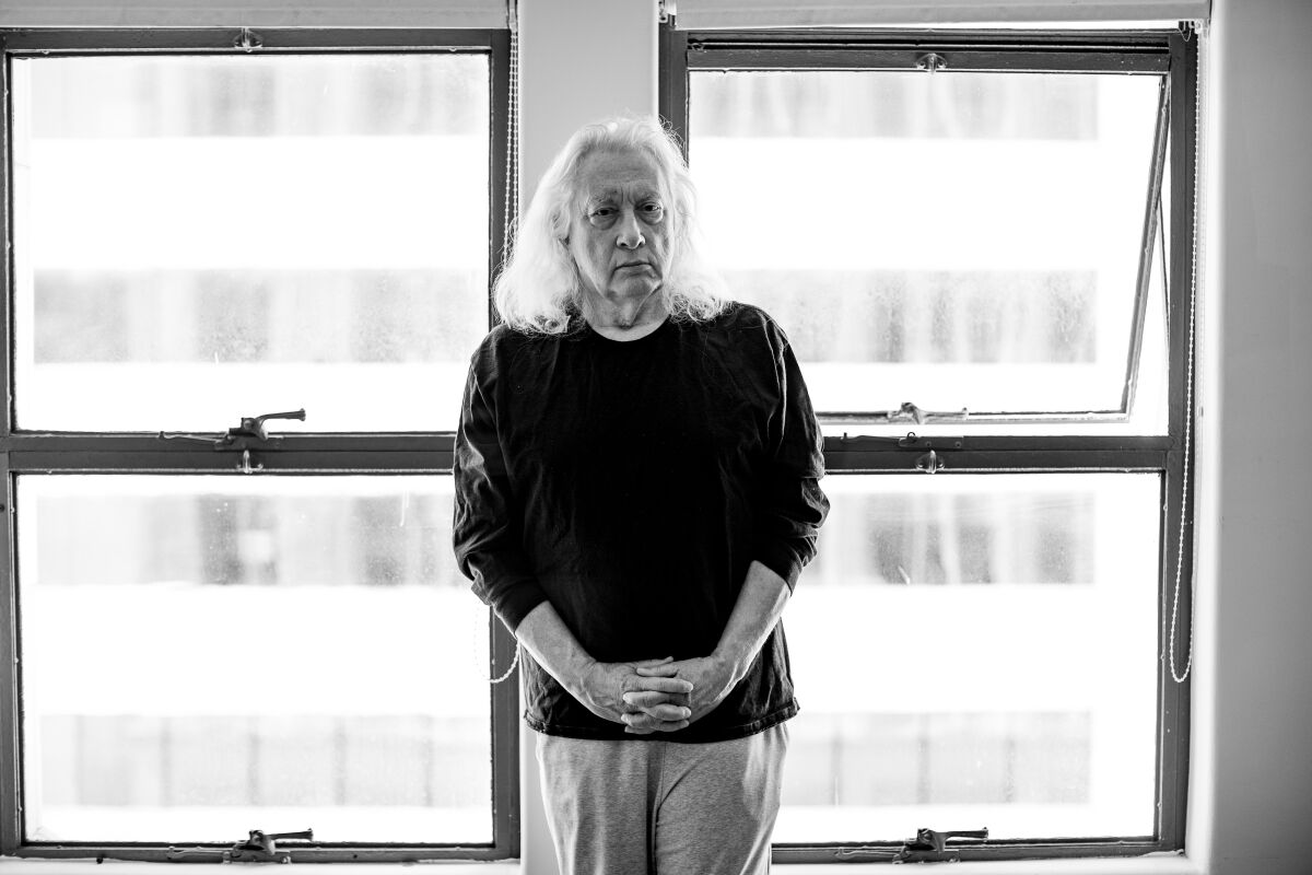 A black-and-white photo of a man with long white hair standing near windows