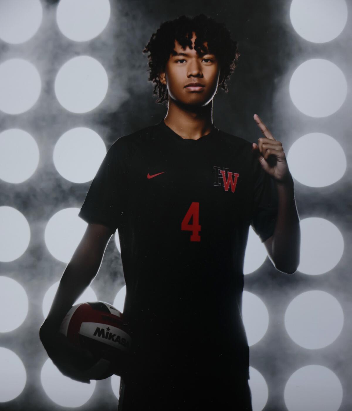  A teenage boy in dark T-shirt with a red No. 4 and the letter W points with one finger and holds a volleyball 