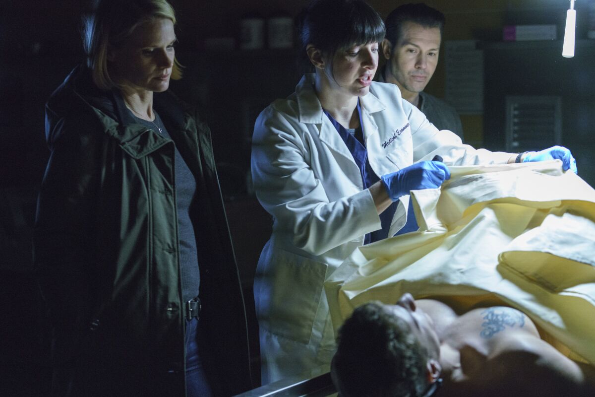 A woman wearing a white lab coat and gloves raises the cover over a body as two others watch.