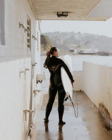 A person in a wetsuit holding a surfboard rinses off in an outdoor shower at the beach.