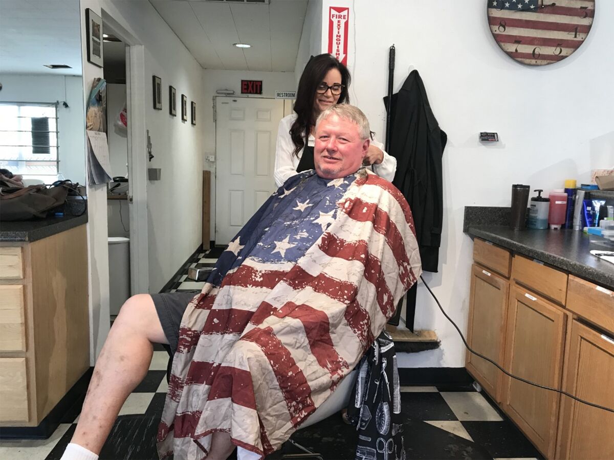 Oildale, Calif., barber Chris Vaughn, in the red Trump hat, finishes up a shave for a regular customer. In the background, the Robert S. Mueller III hearings are on TV.