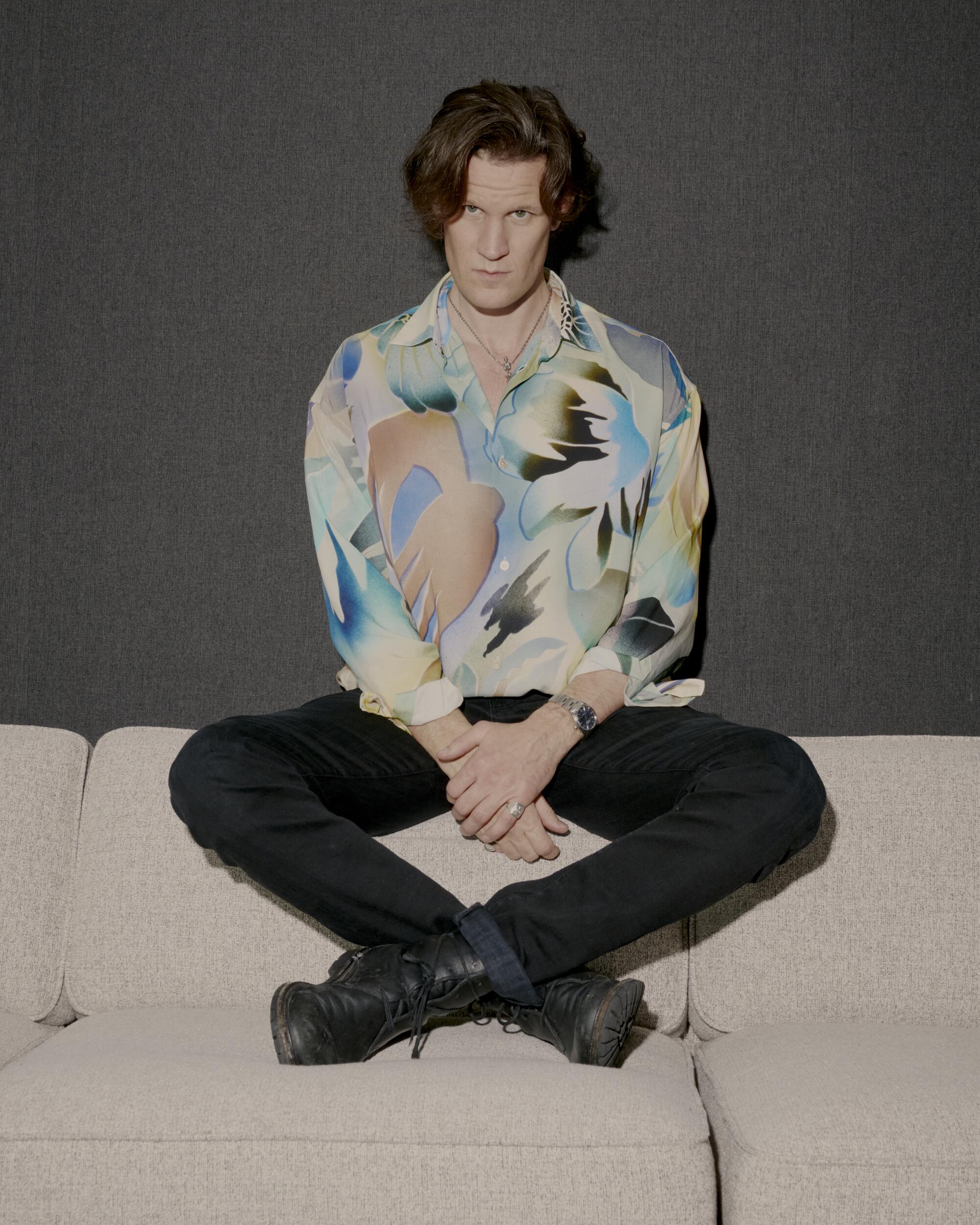 A man in black jeans and a colorful patterned shirt sits on the back of a sofa. 