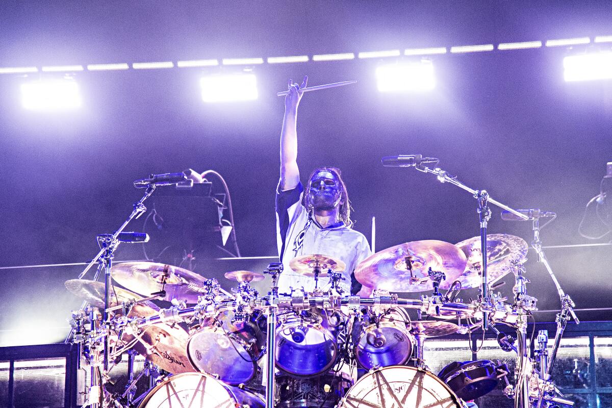 A man wearing a scary mask sitting behind a drum kit on a stage holds up a drumstick in his right hand