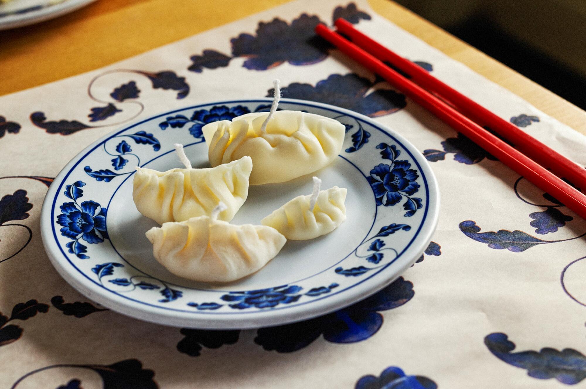 Four candles by Woon in the shape of dumplings rest on a white and blue plate with chopsticks alongside