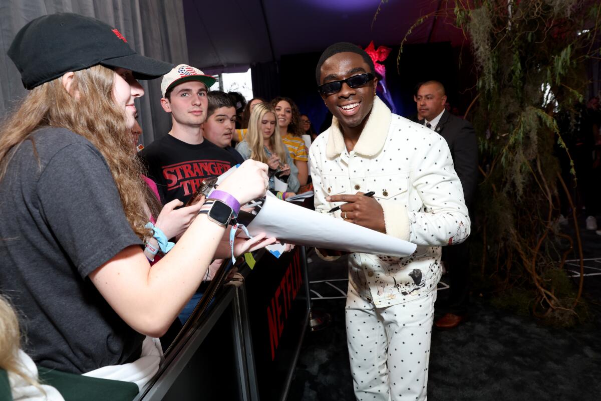 A man in a stylish outfit and shades signs an autograph amid a group of fans