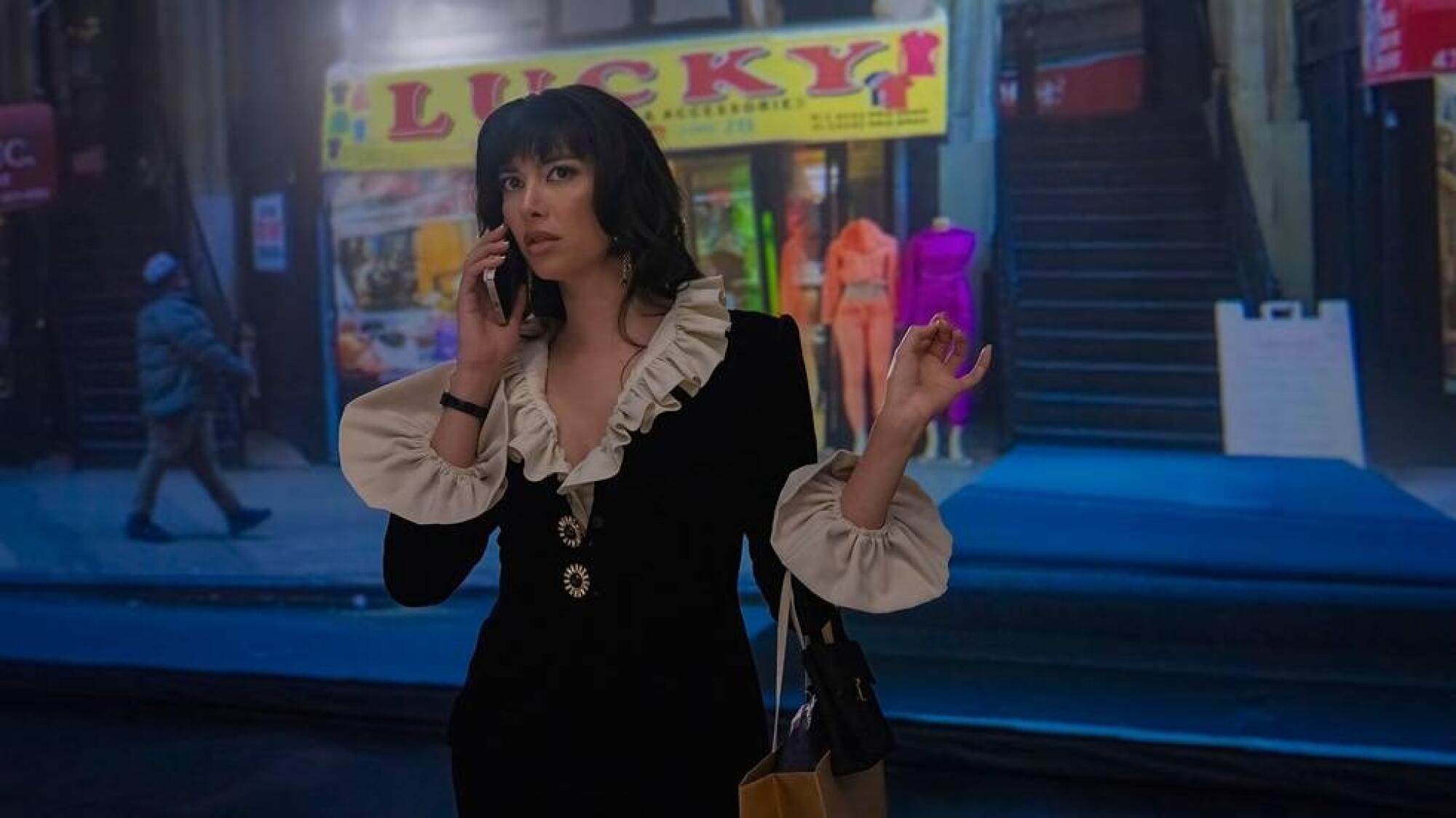 A woman in a black jacket and a white ruffled blouse stands in front of a store with a phone to her ear.