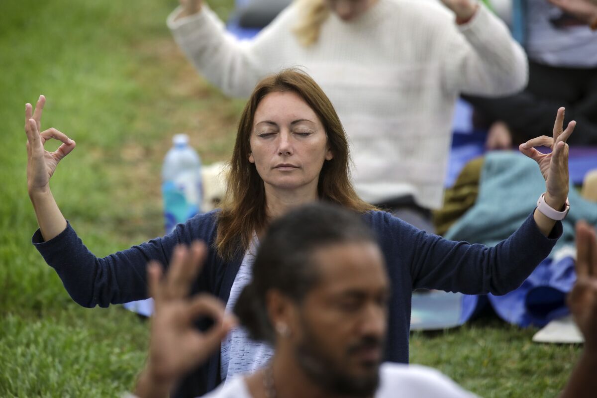 A woman sits in the grass with her eyes closed and hands raised up