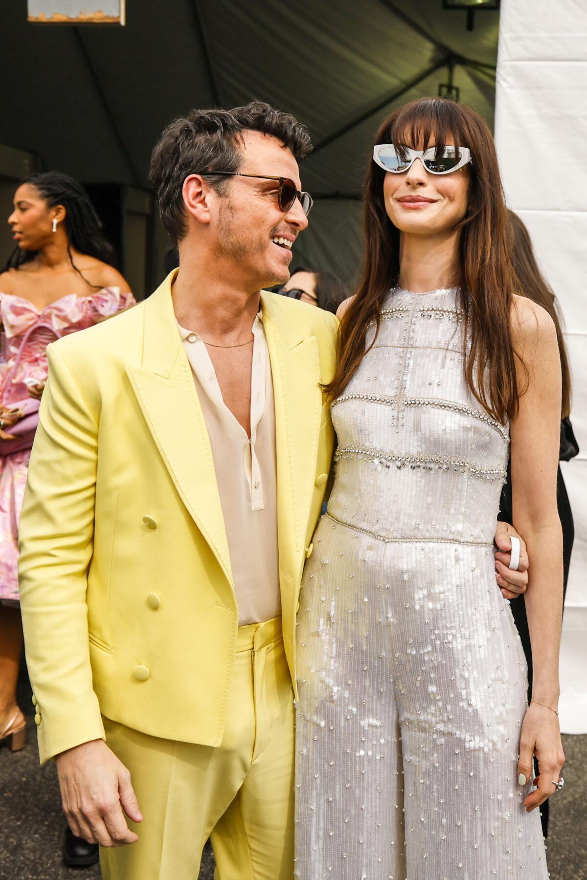 Andrew Scott, in a yellow suit, smiles standing next to Anne Hathaway in a sparkly white jumpsuit