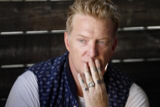 Musician Josh Homme in a printed vest and white t-shirt holds his tattooed fingers in front of his mouth