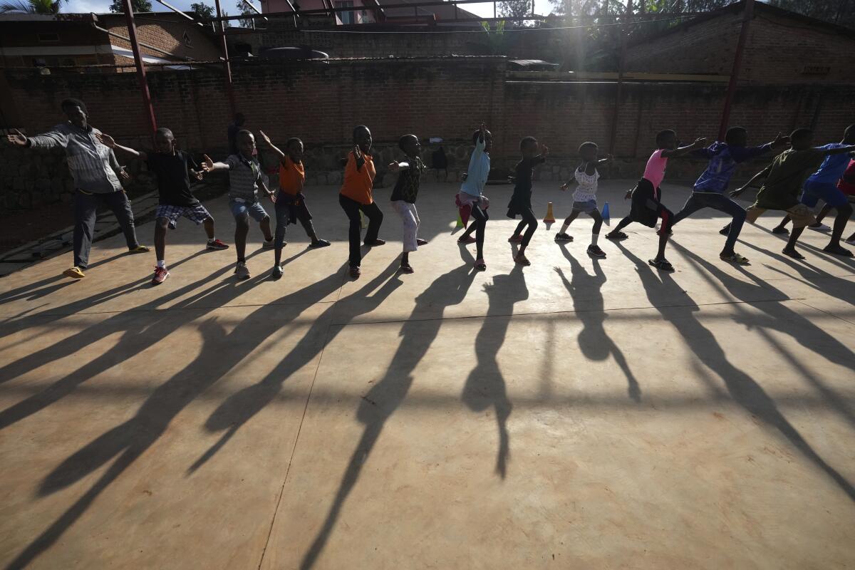 A line of 13 children of various sizes in a fencing stance, arm out, casting long shadows on the pavement in an enclosed area
