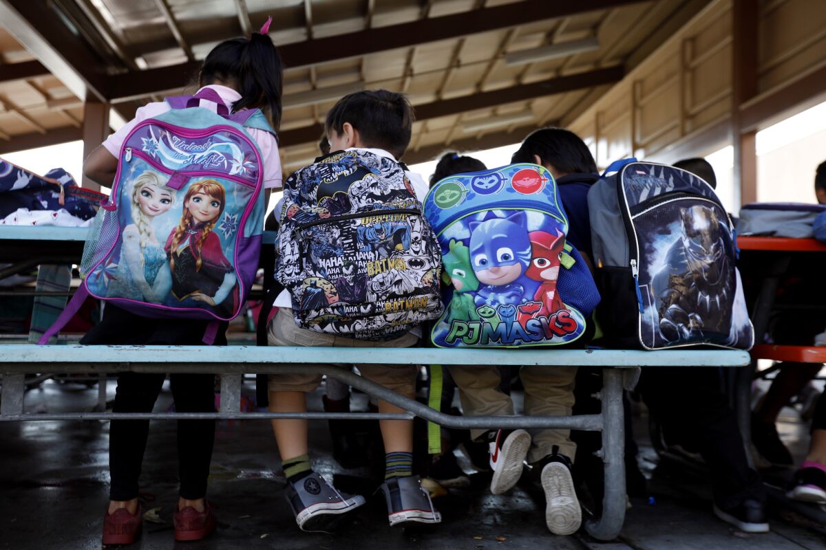 Students sit at lunch tables wearing  backpacks.