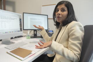 A woman with dark hair, in a cream-colored jacket, gestures with her hands while seated in front of two computer screens