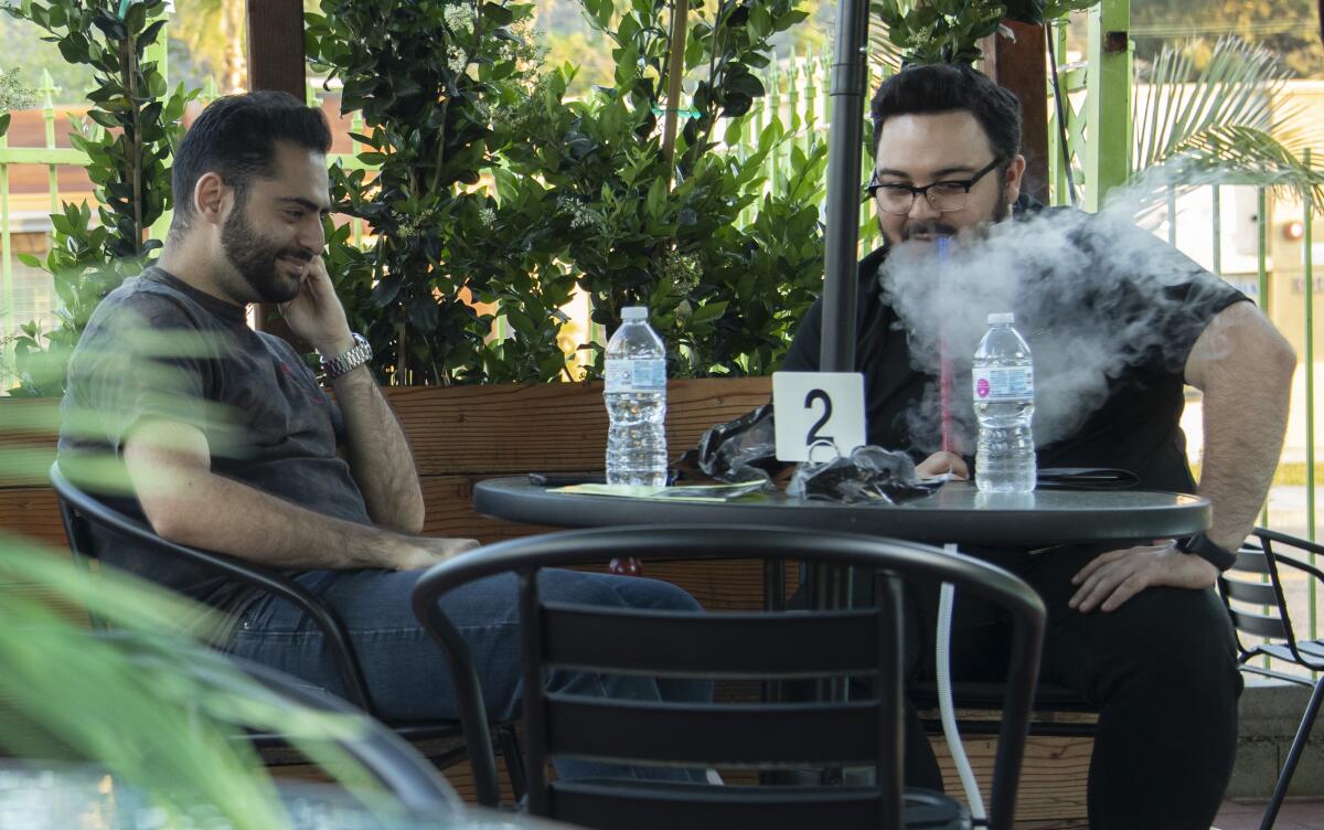 Two men sit at an outdoor table, one of them smoking from a hookah pipe
