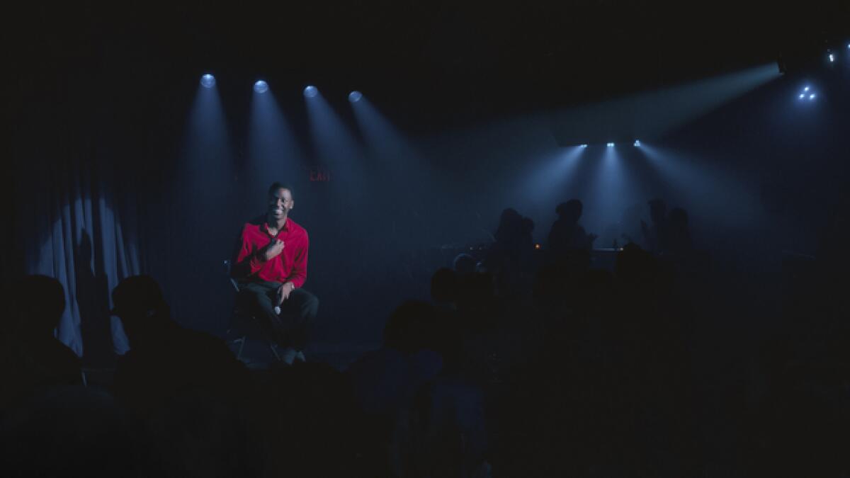 A man in a red shirt sits on a spot-lighted stage