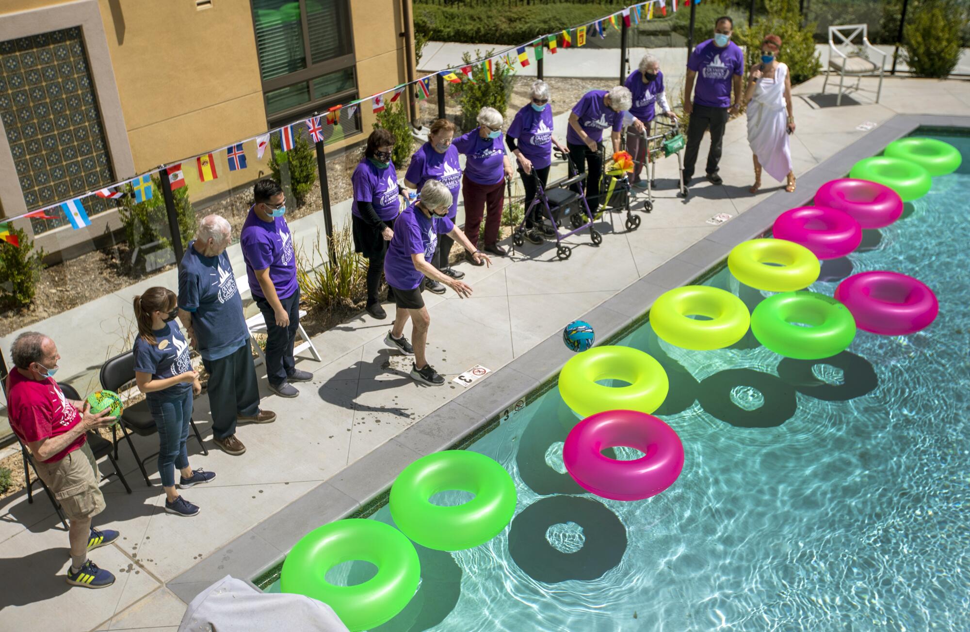 People, many in matching purple shirts, line a swimming pool filled with colorful flotation rings