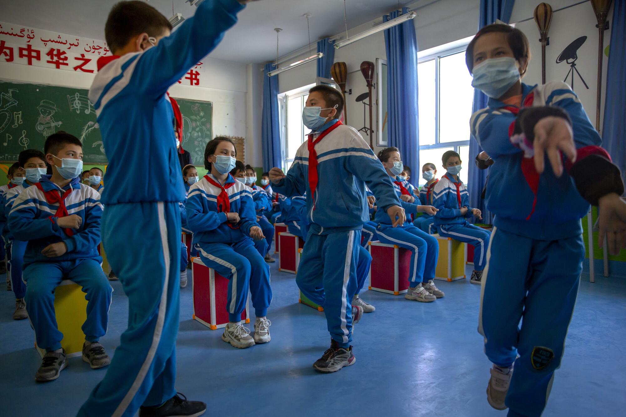 Schoolchildren in light blue and white uniforms with red scarves dance in a classroom 